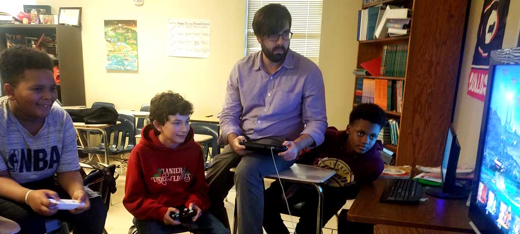 An adult plays video games with two kids while a third youth looks on