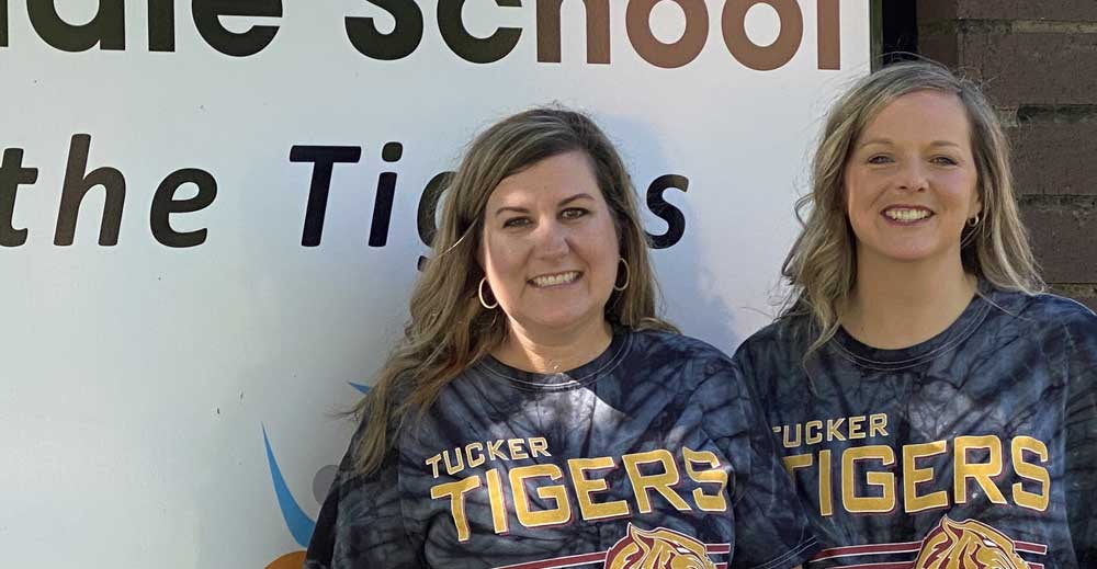 J Lenac and Meredith Moseley wearing matching Tucker Tigers shirts, stand in front of school signage outside