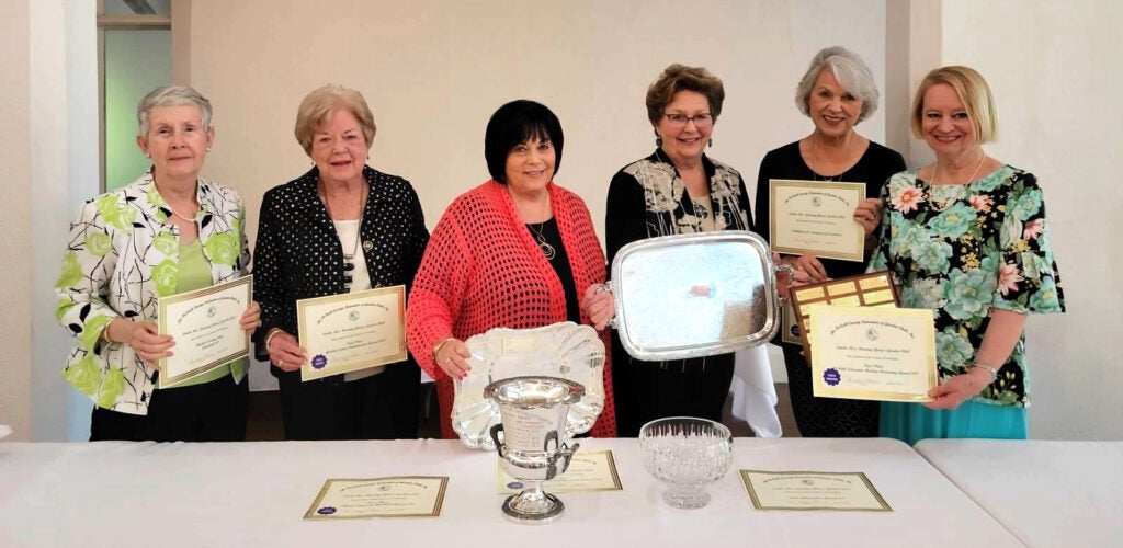 A row of women stand each holding certificates and two holding silver platters. More certificates, a large silver cup, and a crystals dish are on a table in front of them