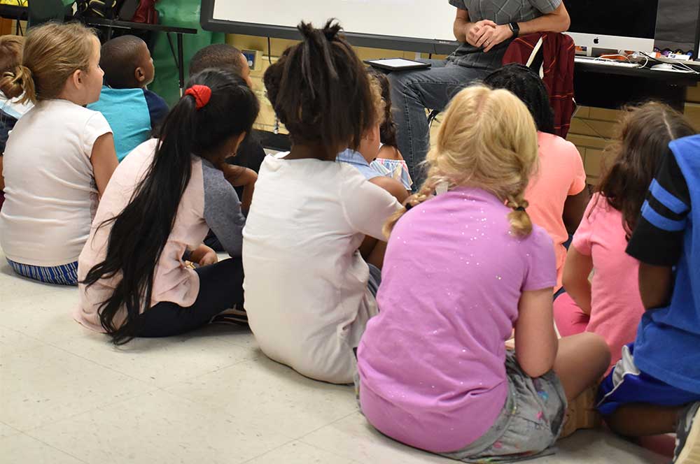 Students sit on floor in front of teacher in chair