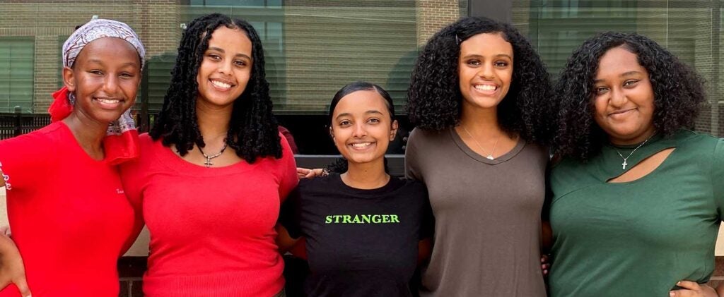 Five girls smile in front of large window