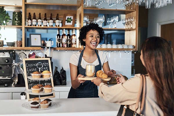 Happy woman, barista and serving customer at cafe for service, payment or order on counter at coffee shop. African person, waitress or employee in small business restaurant helping client at checkout.