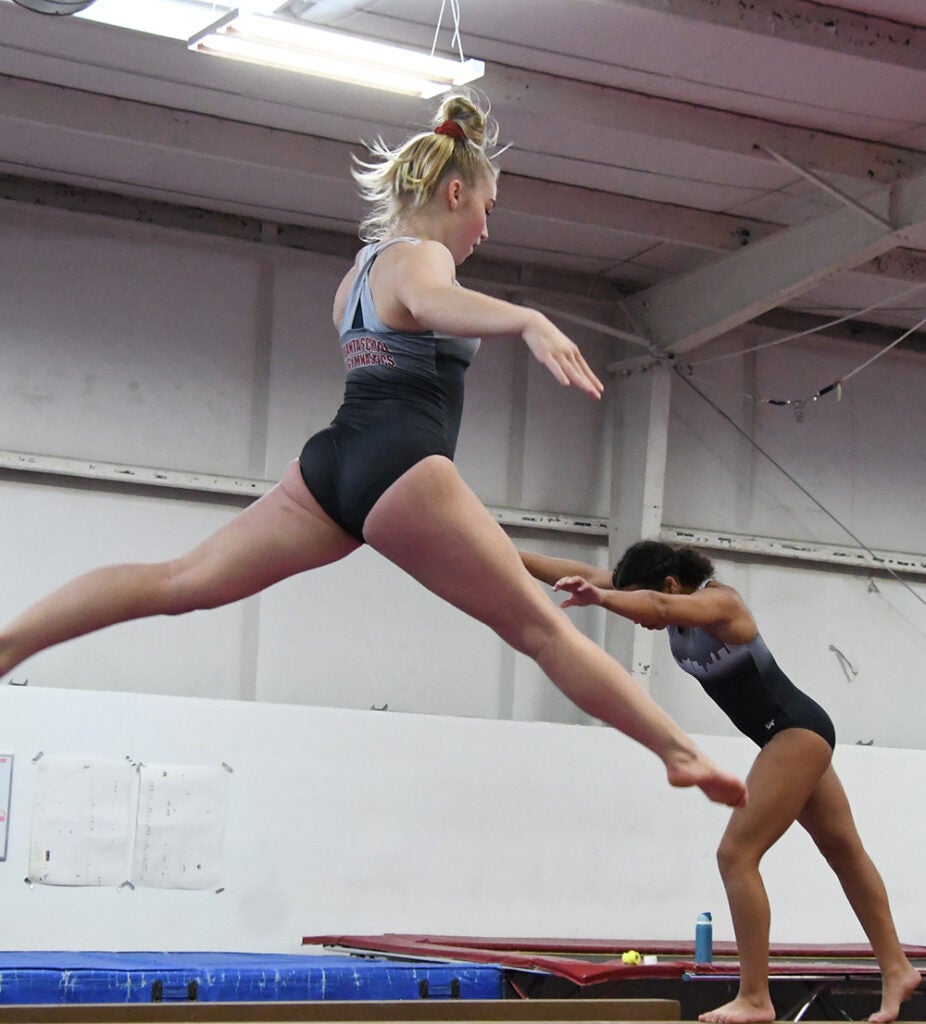 A gymnast jump-splits in the air atop a balance beam. Another gymnast prepares for a cartwheel on another beam in the background.