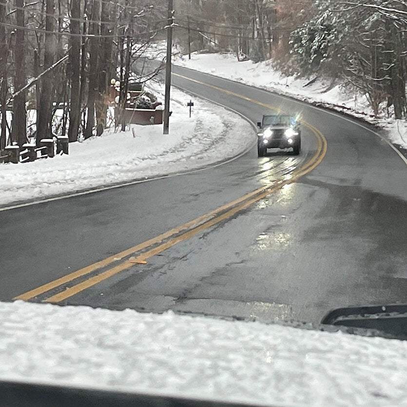 A car drives on a clear road with snow on the surrounding ground and trees.